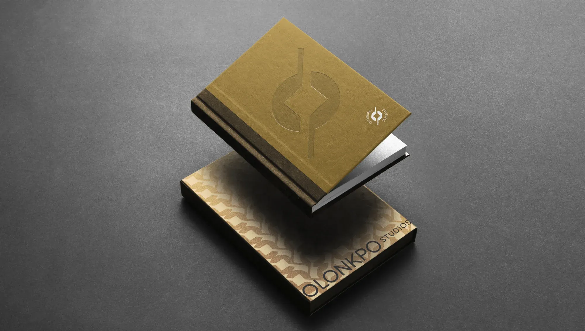 Two gold-foiled notebooks floating above a grey surface, the mark debossed on the cover.